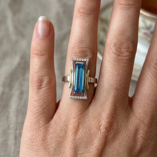 A close-up of a hand wearing a silver and yellow gold ring with a rectangular blue gemstone and small clear accent stones.