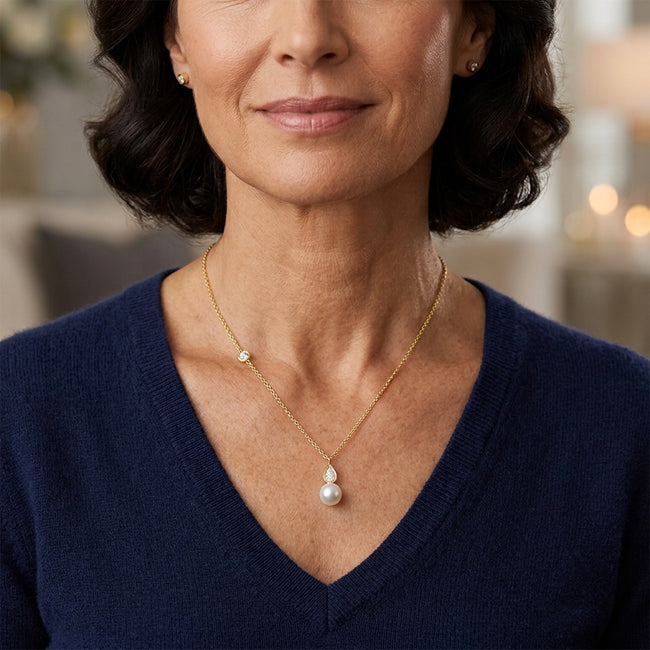 A close-up of a woman wearing a delicate gold necklace with a teardrop-shaped white pearl pendant and a small diamond accent, paired with a navy blue V-neck sweater.